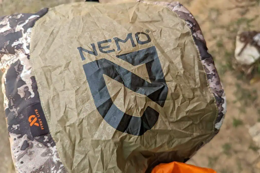 Close-up of a crumpled, camouflage-patterned NEMO Fillo Elite Ultralight Backpacking Pillow bag, with the black NEMO logo prominently displayed. The bag is being held outdoors, with grass visible in the background.