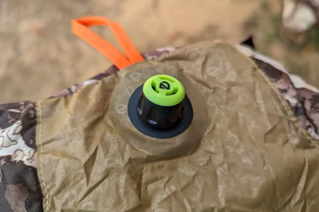 Close-up of an air valve with a green and black cap on a brown, camouflage-patterned NEMO Fillo Elite Ultralight Backpacking Pillow. An orange fabric loop is attached nearby, and the background is out of focus.