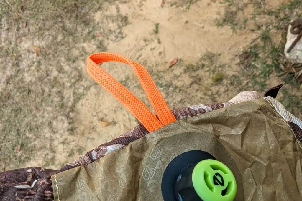 Close-up of a brown and tan outdoor fabric on the NEMO Fillo Elite Ultralight Backpacking Pillow, featuring a bright orange woven loop and a green and black plastic valve, set against a sandy and grassy background.