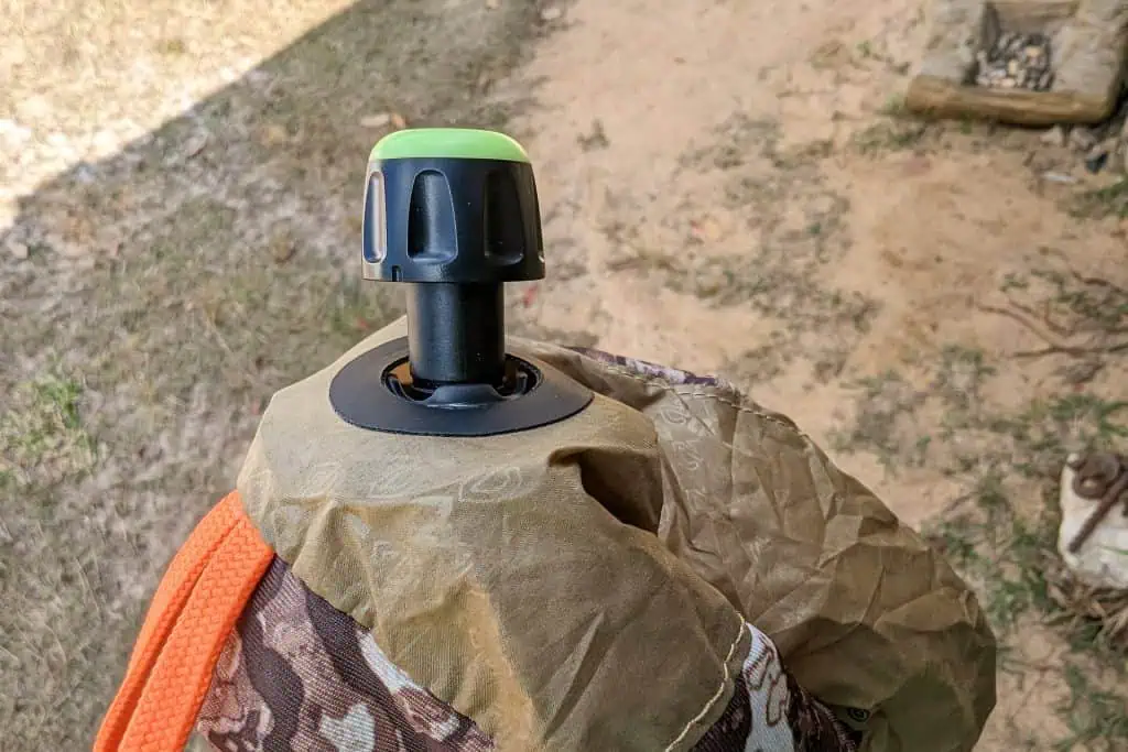 A close-up of a black and green plastic valve on a NEMO Fillo Elite Ultralight Backpacking Pillow, attached to crumpled, camouflage-patterned fabric, with dirt and grass in the background.