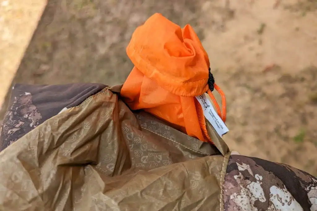 A close-up of an orange drawstring bag, perfect for storing your NEMO Fillo Elite Ultralight Backpacking Pillow, rests on a camouflage-patterned surface outdoors. A product tag is attached, with blurred grass and dirt in the background.