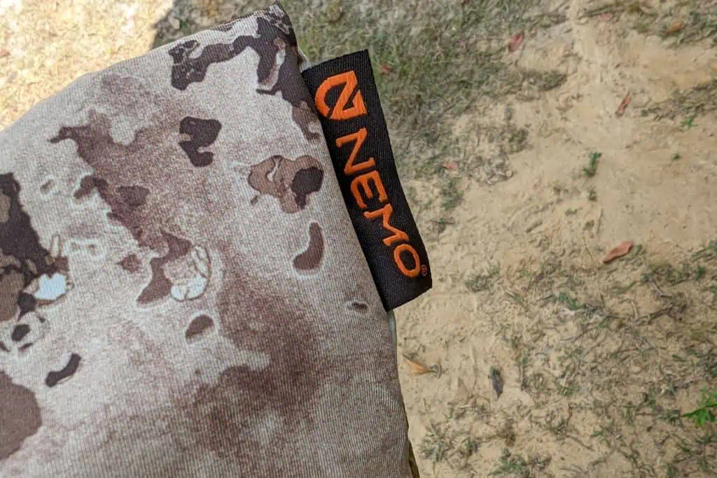 A close-up of camouflaged fabric with an orange and black tag reading NEMO, possibly from the NEMO Fillo Elite Ultralight Backpacking Pillow. The ground in the background is a mix of dirt, grass, and scattered leaves.