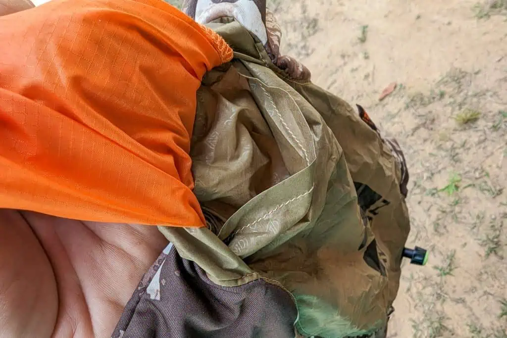 A close-up of a hand holding a crumpled orange and green nylon fabric, possibly outdoor gear like the NEMO Fillo Elite Ultralight Backpacking Pillow, with sand and sparse grass visible in the background.