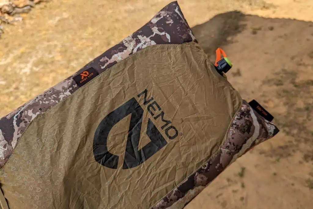 A close-up of a NEMO Fillo Elite Ultralight Backpacking Pillow with a camouflage pattern, lying outdoors on dry ground, showing the logo and an orange valve on the right side.