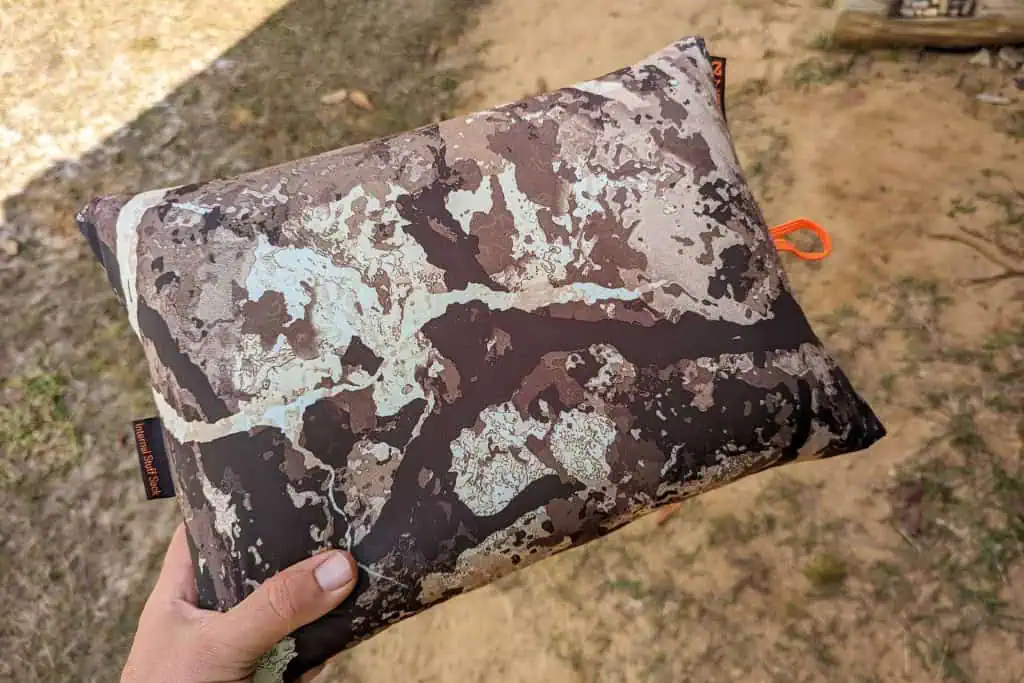 A hand is holding a rectangular pouch with a brown and tan camouflage pattern, revealing the compact design of the NEMO Fillo Elite Ultralight Backpacking Pillow. Sandy ground with patches of grass completes the outdoor backdrop.
