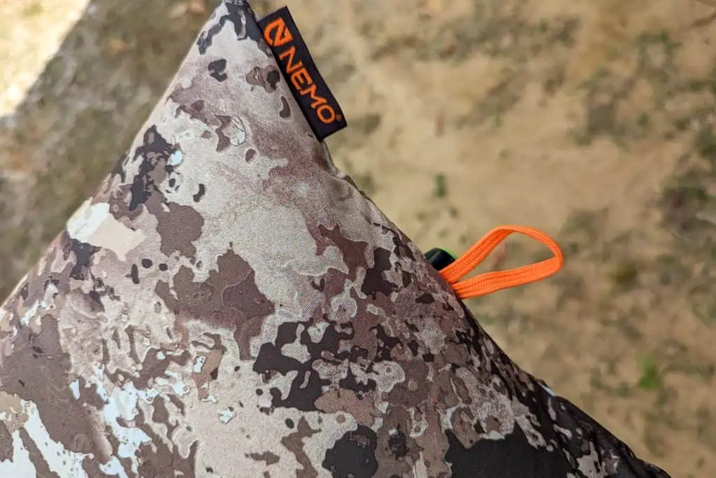 A close-up of a camo-patterned NEMO Fillo Elite Ultralight Backpacking Pillow with an orange loop, set against a blurred dirt and grass background.