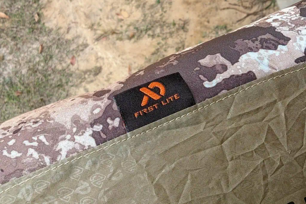 A close-up of a First Lite brand tag on camouflage-patterned outdoor fabric, with a NEMO Fillo Elite Ultralight Backpacking Pillow peeking out and grass and dirt visible in the background.