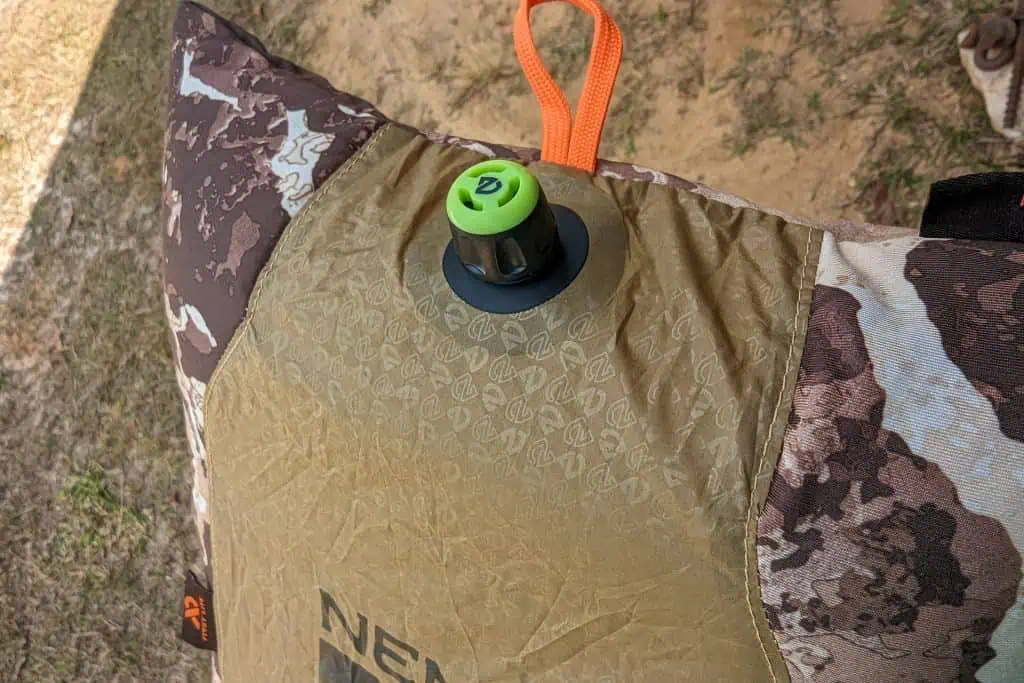 Close-up of a NEMO Fillo Elite Ultralight Backpacking Pillow in brown and beige camo, featuring an orange loop and green-and-black air valve, shown outdoors on grass and dirt.