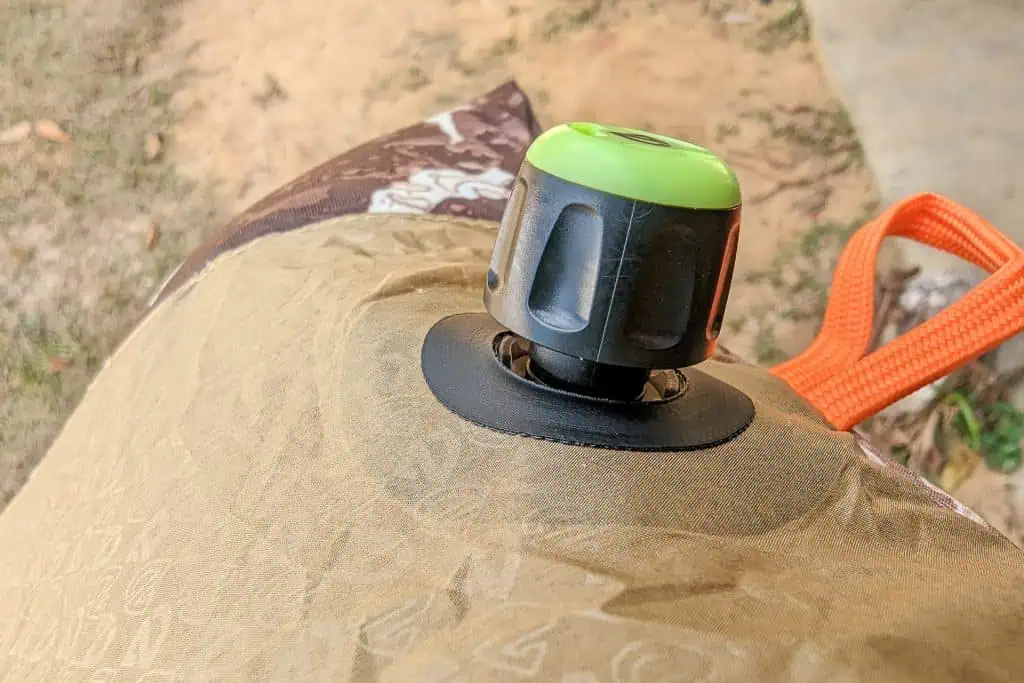 Close-up of a green and black valve on a tan inflatable outdoor cushion or mattress, likely the NEMO Fillo Elite Ultralight Backpacking Pillow, with an orange carrying handle and dirt ground in the background.